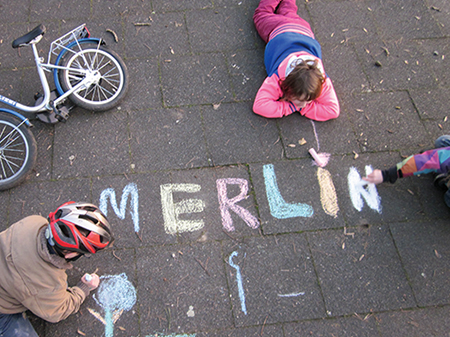 Kinder schreiben mit bunter Kreide das Wort "Merlin" auf den Asphalt.
